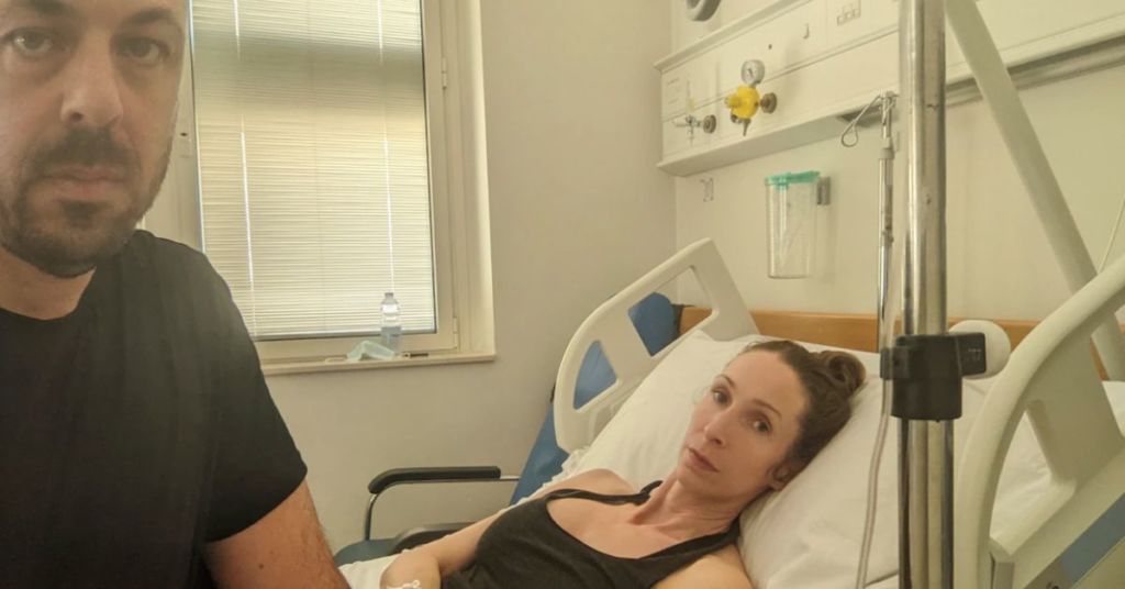 Andrea Prudente, 38, who suffered an incomplete miscarriage while vacationing in Malta, lies in bed at the Mater Dei Hospital in Msida, Malta, comforted by her partner Jay Weeldreyer, left, in June 2022. (Jay Weeldreyer/AP) 